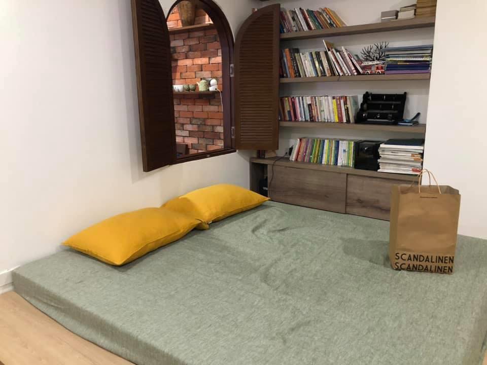 Asparagus green yarn-dyed French linen fitted sheet with mustard yellow pillowcases on a floor mattress in a Hanoi old quarter bedroom with arched window and bookshelf