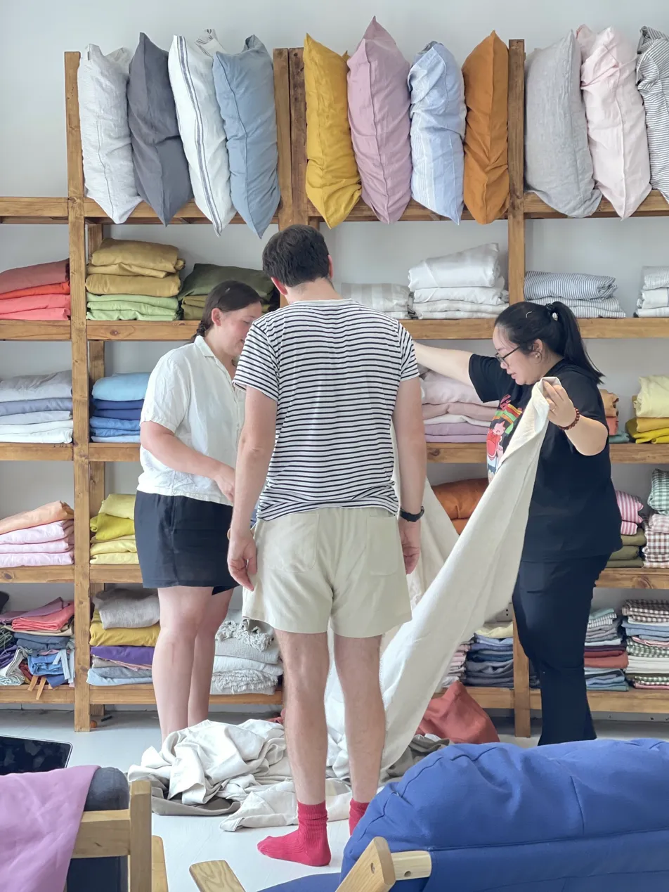 International customers browsing SCANDALINEN linen bedding workshop in Hanoi Vietnam with full colour range displayed on wooden shelves